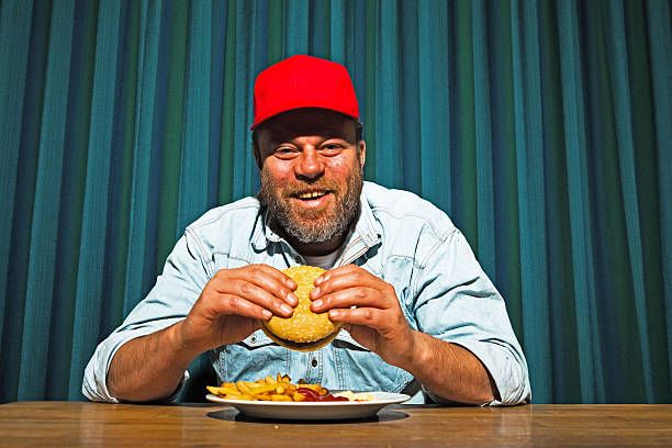 Man with beard eating fast food meal. Enjoying french fries and a hamburger. Trucker with red cap.
