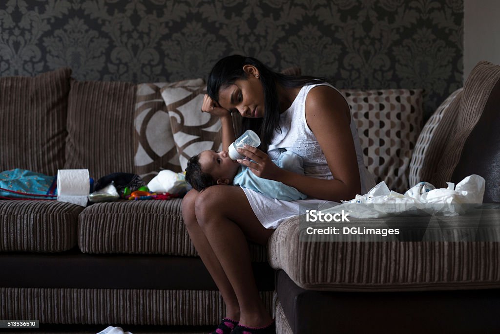 Give me break! Stressed young mother sitting on her sofa whilst feeding her baby son. She has her head in her hand and is surrounded by mess. Mother Stock Photo