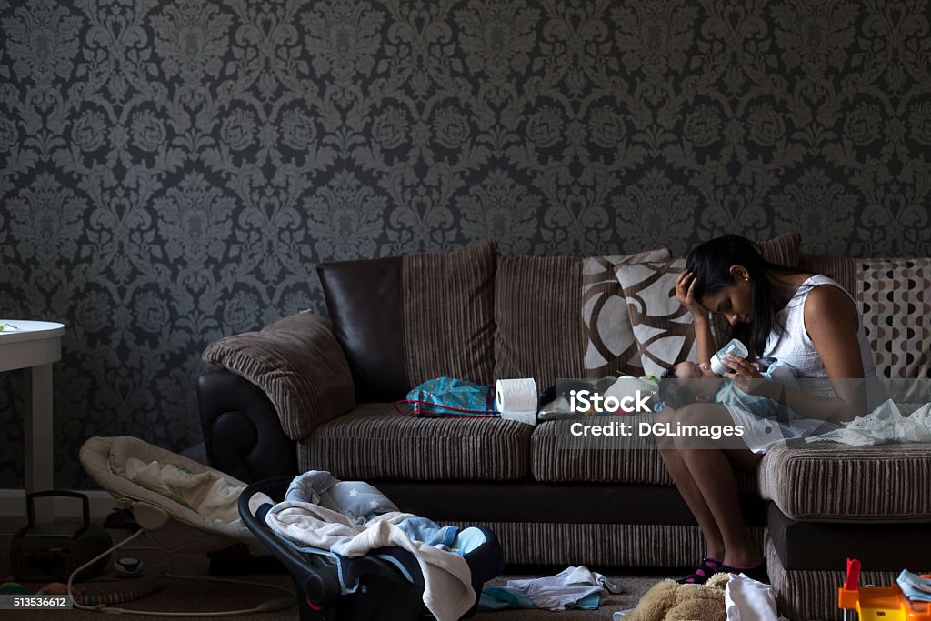 Give me break! Stressed young mother sitting on her sofa whilst feeding her baby son. She has her head in her hand and is surrounded by mess. Chaos Stock Photo