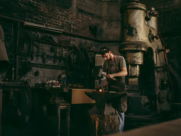 Portrait of a caucasian young adult craftsman hammering iron on an anvil in a blacksmith's fabric