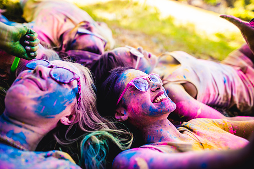 Multi-Ethnic group covered in colorful powder lay in grass laughing in a park at a Holi festival in the summer