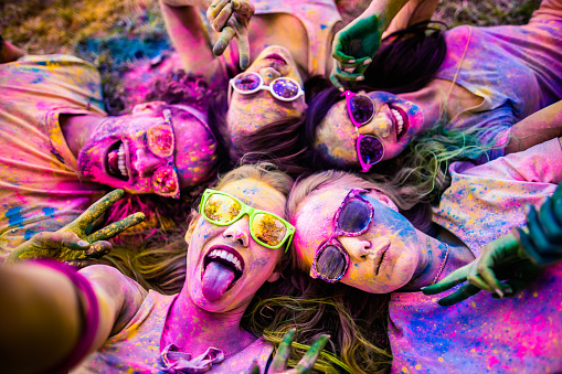 Multi-Ethnic group covered in colorful powder lay in grass taking a selfie in a park at a Holi festival in the summer