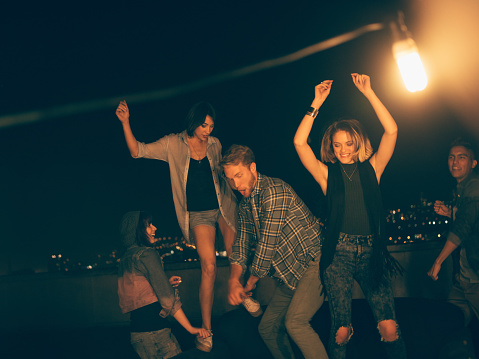 Multi-ethnic group of teenager friends dancing with raised arms and laughing during a night rooftop party