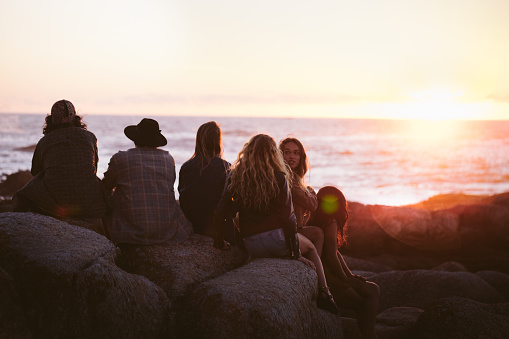 Group of young adult friends enjoying a sunset at the sea sitting on rocks