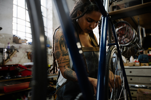 Focused female bicycle mechanic with tattoos on her arm looking serious and focused while working on a bike in her repair shop