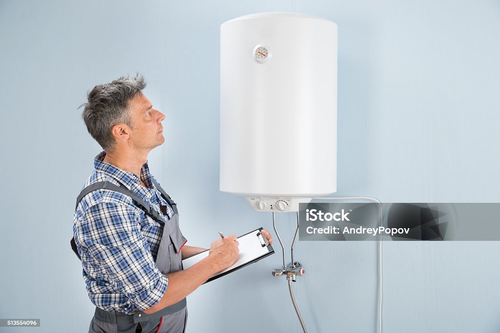 Male Plumber With Clipboard Looking At Electric Boiler Portrait Of Mid-adult Male Plumber Holding Clipboard Looking At Electric Boiler Water Stock Photo