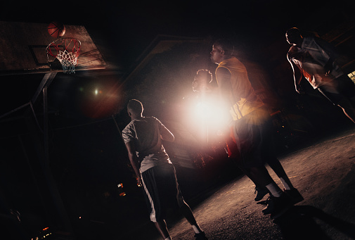 African American basketball players watch ball go into basket in inner-city court during nighttime with single light in background