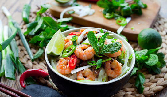 Vietnamese prawn noodle , Tasty traditional  with rice noodles, spring onions, king prawns, bird's eye green, red chilies and shiitake mushrooms. Decorated with piece of fresh lime, leaves of mint, basil and slices of spicy peppers. All in bowl with chopsticks on natural wooden background.
