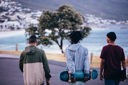 Multi-Ethnic group of skater friends walking down street along seaside, carrying skateboards