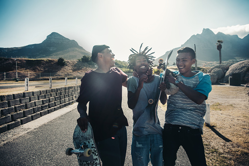 Multi-Ethnic Group of Skater friends standing together, holding skateboards and laughing out loud on road at seaside with sun shining behind them