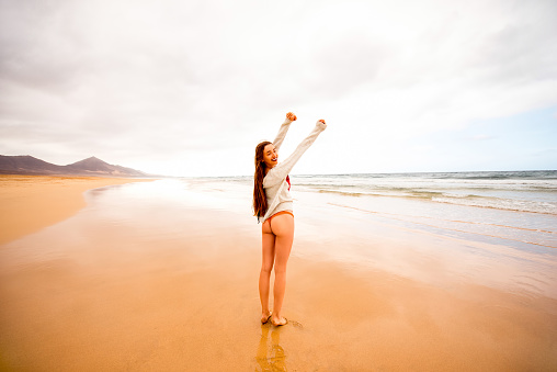 Young woman in sweater having fun enjoying beautiful sandy beach on the foggy weather on Fuerteventura island in Spain. General plan with a lot of space