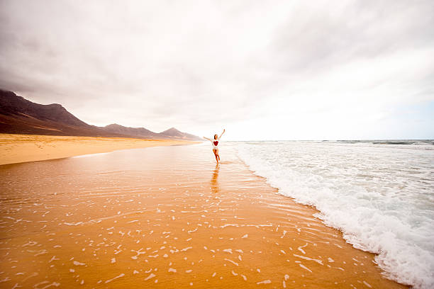 Young woman in sweater having fun enjoying beautiful sandy beach on the foggy weather on Fuerteventura island in Spain. General plan with a lot of space