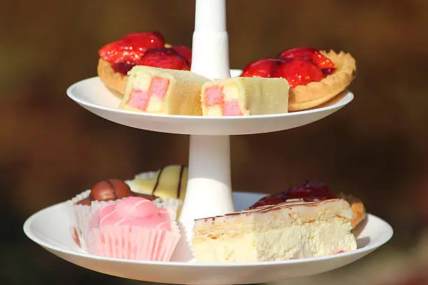 Photo showing a white cake stand with tiers, pictured in the garden and set for a traditional English afternoon tea.  On these top tiers are cakes and sweet nibbles, including mini Battenbergs, fresh cream slices, fondant fancies (French fancy cakes) and strawberry tarts.