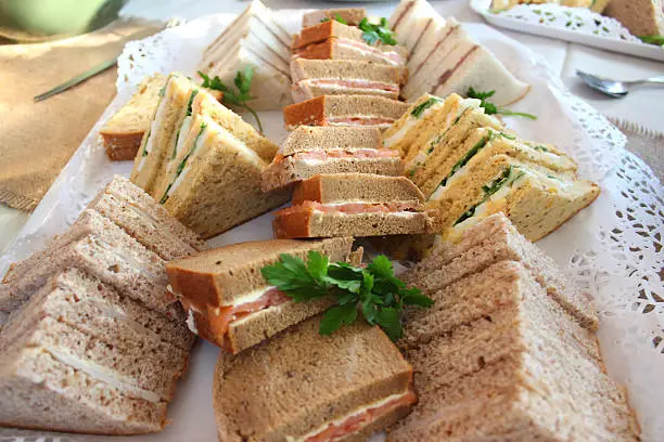 Photo showing a sandwich platter made using freshly baked brown granary bread and white bread.  The triangular sandwiches are being served as part of a party food / finger-food buffet and include ham, cheese, smoked salmon and cream cheese, beef and horseradish, and egg salad, cress and lettuce - looking rather like they could be served as part of a traditional afternoon tea.