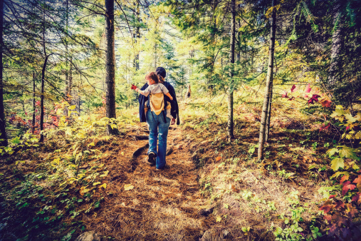 A mother is hiking on a trail in a forest with her baby in a back carrier during the autumn season.  Filtered to give retro, faded look.