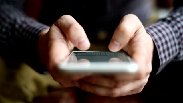 man typing a message on a smartphone. face is not visible. man wearing a plaid shirt and jeans. reflected on the smartphone screen man's fingers.  close-up. open aperture. average lighting. one person