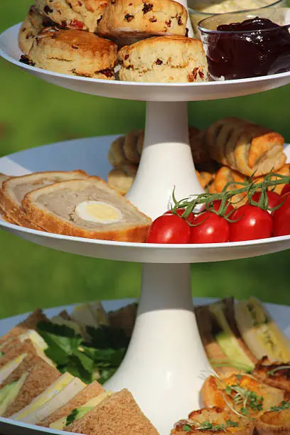 Photo showing a white cake stand with tiers, pictured in the garden and set for a traditional English afternoon tea.  On the bottom tier are egg and cress sandwiches (white bread) and cheese and cucumber sandwiches (brown bread), as well as some homemade mini quiches / egg flans.