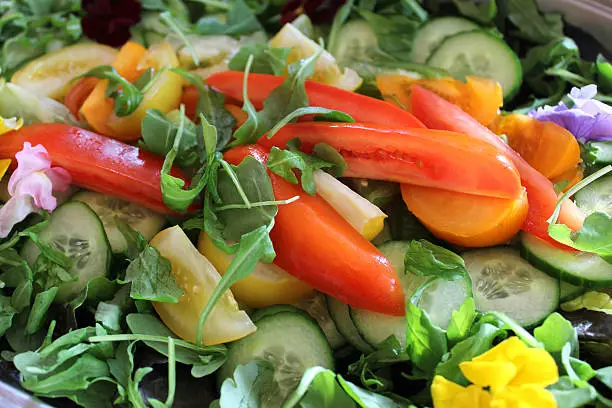 Photo showing a large bowl of green salad, made with lettuce leaves, rocket leaves, slices of cucumber and chunks of homegrown red and yellow tomatoes.  The dish is being served as part of a party food buffet and is pictured garnished with edible flowers (yellow and purple pansies and nasturtiums).