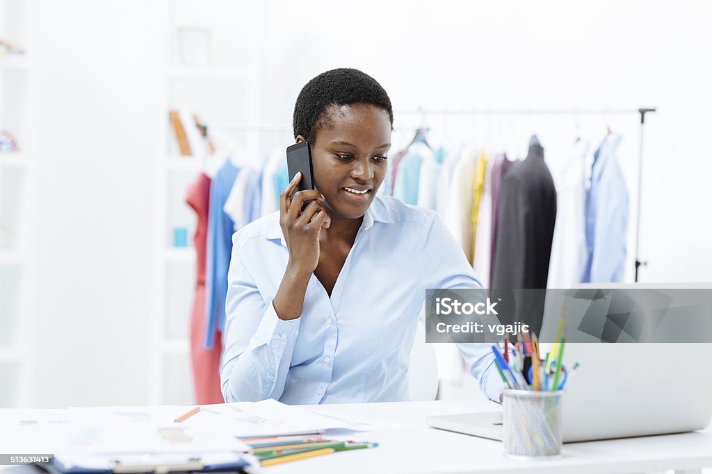 Female fashion designer in the office Portrait of an african woman, fashion designer. Sitting at her desk, talking on the phone and looking at laptop. 30-39 Years Stock Photo