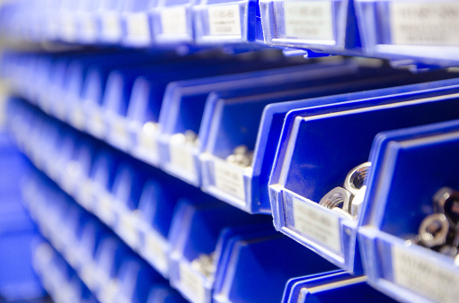 Rows of blue plastic storage bins containing stocks of parts and components, such as screws, bolts and nuts, in an industrial or commercial warehouse shot with selective focus