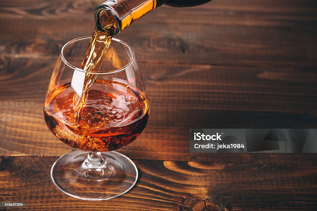 Pouring cognac or whiskey from the bottle Pouring cognac or whiskey from the bottle into the glass against wooden background Activity Stock Photo