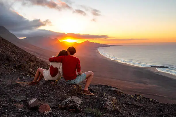 Young couple enjoying beautiful sunset sitting together on the mountain with great view on Cofete coastline on Fuerteventura island