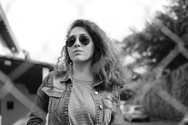 Look through fence of a young urban lady posing as rebel, modern and careless on the street. She is wearing sunglasses, t-shirt and denim jacket. She has curly hair. Monochrome style.