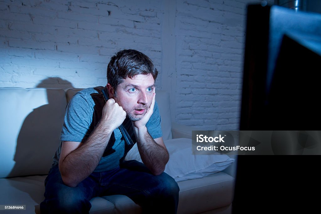 television addict man watching tv holding remote control mesmerized young attractive man at home lying on couch at living room watching tv holding remote control looking mesmerized and intense in television addict concept Television Set Stock Photo