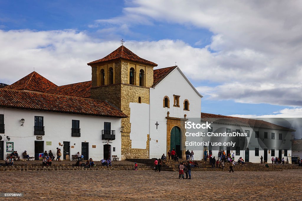 Villa de Leyva, Colombia: Church and people; 16th Century plaza Villa de Leyva, Colombia - September 13, 2014: Founded in 1572 and located at just over 7000 feet above sea level on the Andes Mountains, Villa de Leyva was declared a National Monument in 1954 to protect it's colonial architecture and heritage. It is located in the Department of Boyaca, in the South American country of Colombia.  Photo shows the church on the main plaza or square.  People visiting the town and local residents are seen on the square just sitting and relaxing, chatting with friends or going about their routine chores. Photo shot in the late afternoon sunlight; horizontal format. Copy space. 16th Century Style Stock Photo