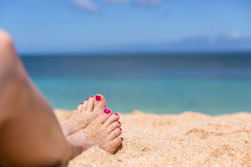 The close-up photograph of a woman's feet on the beach. The background of the blue sea and sky is blurred whilst the woman's feet are in clear focus. The two feet are covered in yellow sand and the toes are painted with a crimson pink nail polish.
