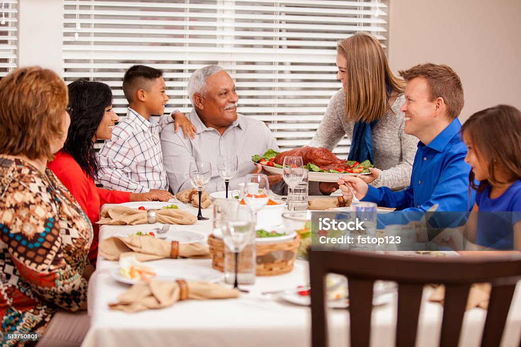 Multi-ethnic, generation family enjoys thanksgiving dinner. Hispanic, caucasian family prepares to enjoy holiday dinner together. Daughter-in-law serves turkey to family.  Dining table. Grandparents, children, grandchildren.  Thanksgiving - Holiday Stock Photo