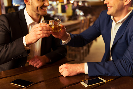 Two young businessmen are smiling and clanging glasses of  alcoholic beverage together while sitting in pub