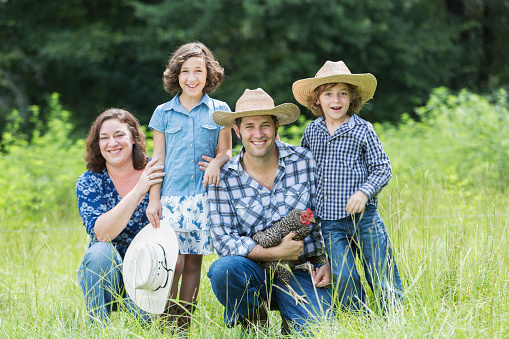A family with two children on a farm, posing in a field, smiling at the camera.  The father is holding a chicken in his arms. They are wearing jeans, cowboy hats and plaid shirts.
