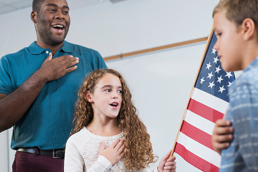 An African American teacher and a multi-ethnic group of elementary school children standing in the classroom saying the pledge of allegiance. The focus is on a 9 year old Hispanic girl who is holding the American flag, hand on heart.
