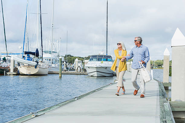 Happy senior couple walking along harbor holding hands A happy, senior couple walking together holding hands along a boat dock, enjoying the water and view of the luxury sailboats on a sunny summer day. They are smiling and relaxed, enjoying retirement. marina stock pictures, royalty-free photos & images