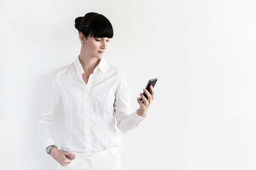 Young confident woman in white clothes, looking like a CEO, is standing at a white wall in an office with a smartphone in her hands. She is holding it in her one hand and looking at it while the other hand is in her pocket. The woman has a tidy hairstyle and overall elegant look. Plenty of copy space available.