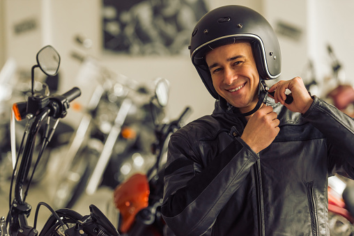 Attractive young blond man in black leather jacket is looking at camera, adjusting his helmet and smiling while standing in a motorbike salon