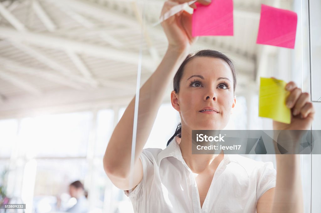 Young businesswoman putting adhesive notes on glass wall in office Beautiful young businesswoman putting adhesive notes on glass wall in office Adhesive Note Stock Photo