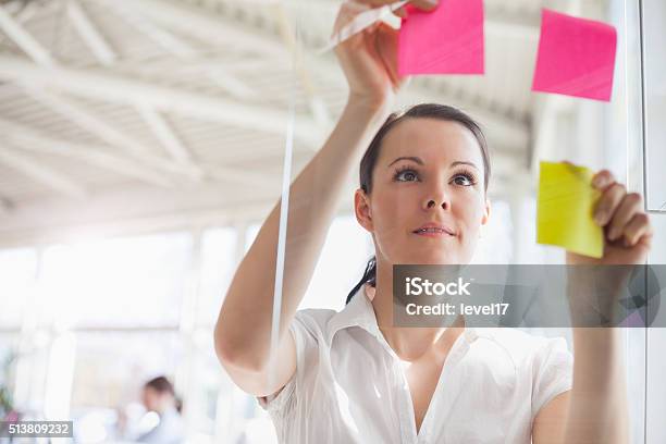 Young Businesswoman Putting Adhesive Notes On Glass Wall In Office Stock Photo - Download Image Now