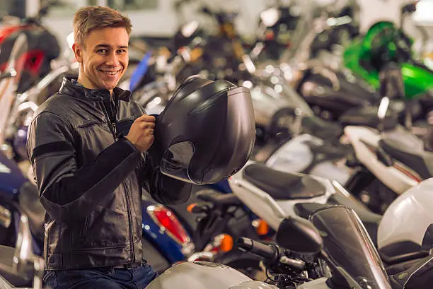 Attractive young blond man in black leather jacket is holding a helmet, looking at camera and smiling while sitting on a motorbike