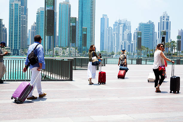 United Arab Emirates, Dubai - May 17, 2014: Capture of caucasian tourists walking with luggage aroud Dubai Mall and mere with fountain. Behind mere is modern skyline with huge residential buildings and hotels.