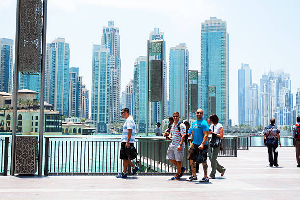United Arab Emirates, Dubai - May 17, 2014: Capture of caucasian tourists outside of Dubai Mall. Group of men is walking along mere and fountain. Behind man a woman is following. At right side are more people. In background is skyline of modern residential buildings and hotels.