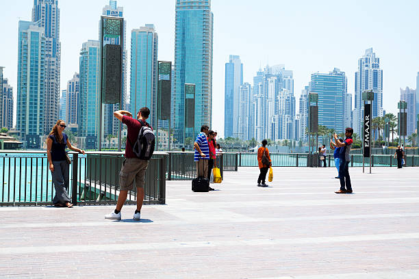 United Arab Emirates, Dubai - May 17, 2014: Scene outside of Duabi Mall. On promenade around mere and fountain are several groups of tourists. Most are taing photos. At left side is caucasian couple, man is taking photo of woman. At right side are indian people, a man is photographing group. In background is modern skyline.