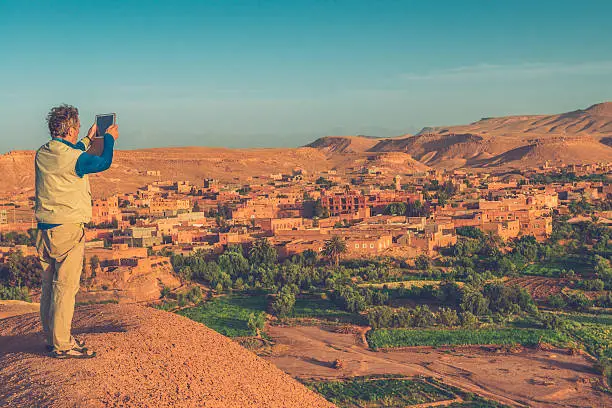 View of senior man from behind while he is standing on the hill and photographing famous old fortified city (ksar) Aït Benhaddou.Located in the foothills on the southern slopes of the High Atlas in the Province of Ouarzazate, in Ounila Valley, central Morocco. This ancient city, giant fortification, is an example of earthen clay architecture. It is made up of six Kasbahs and nearly fifty ksars which are individual Kasbahs. Many films have been shot there.  It is a UNESCO World Heritage Site. Nikon D800, full frame, XXXL.