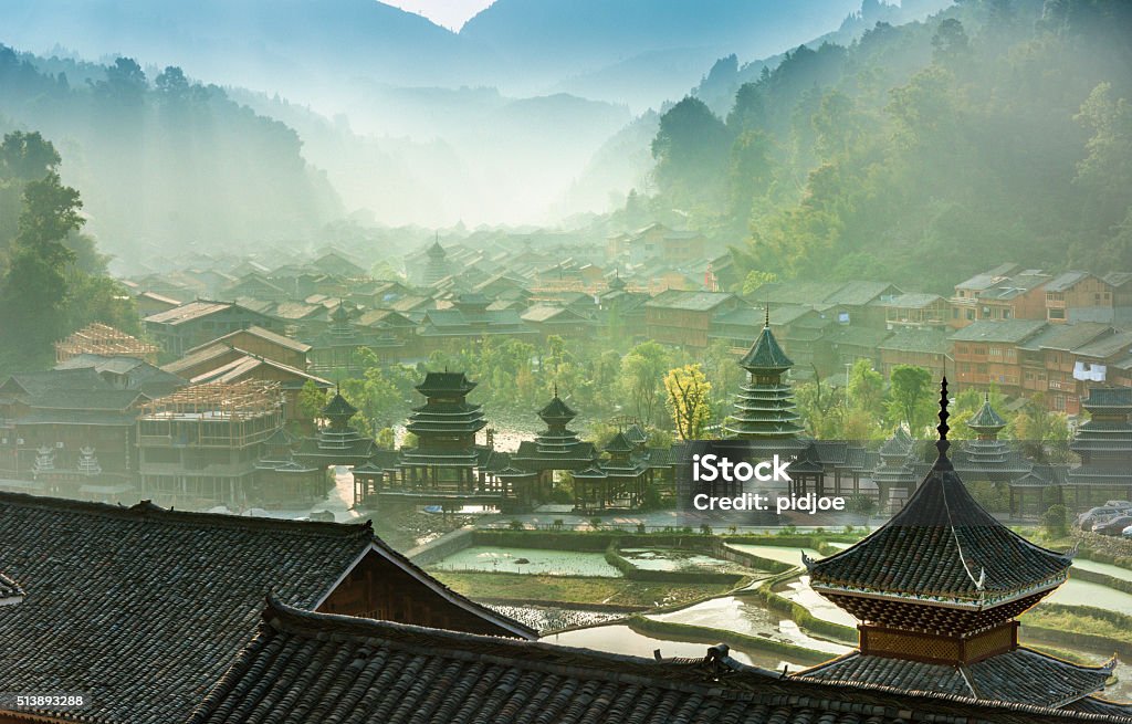 Early morning , Entrance  village Zhao Xing, rain and wind bridge. Sunrise,fog, Rice paddy in the front, village and mountains in the back. Roof of a temple in front. Guizhou Province Stock Photo