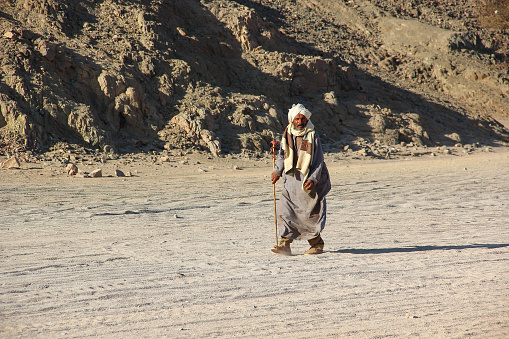 Hurghada, Egypt - April 24 2015: The old Bedouin with a stick walking through the desert on the background sand and mountains, Egypt, Hurghada on April 24, 2015.