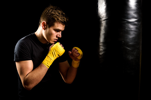 Attractive young man in yellow boxing wraps is training and hitting heavy bag. Fitness, aerobic exercise for active people. Strong athlete isolated on black.