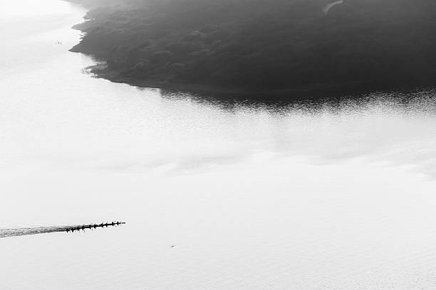 Canoe Paddlers race across dam waters silhouetted birds-eye overhead air landscape in black and white vintage