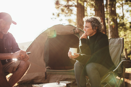 Portrait of relaxed mid adult couple sitting outside tent while camping nearby lakeside on a sunny day. Woman holding coffee talking with man.