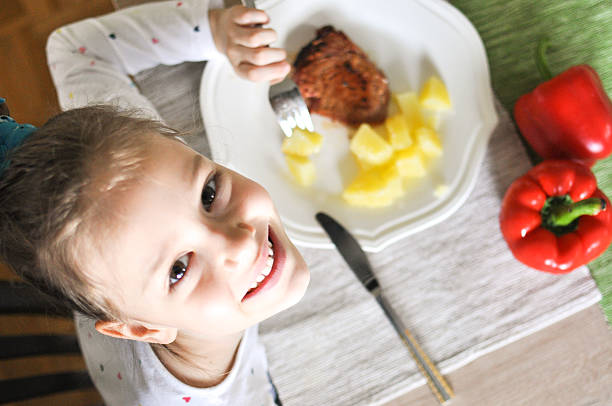 Wanna have lunch with me? Young girl eating grilled tuna steak and boiled potatoes, Indoors shot of a little girl having a healthy meal decorated with red paprika and looking up at camera. girl eating steak stock pictures, royalty-free photos & images